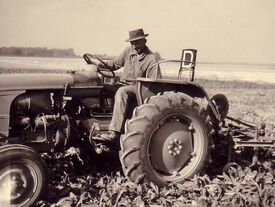 Photo ancienne. Un fermier sur son tracteur tirant une herse précédant une personne qui arrache les mauvaises herbes dans un champ cultivé.  