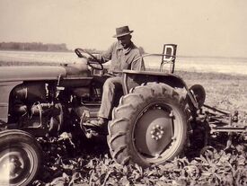 Photo ancienne. Un fermier sur son tracteur tirant une herse précédant une personne qui arrache les mauvaises herbes dans un champ cultivé.